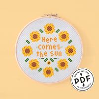 Esquema de punto de cruz para aro de bordar redondo. Frase sobre salud mental "here comes de sun" en letras amarillo oscuro rodeada de girasoles de color amarillo y hojas de color verde.